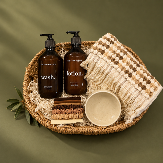 A curated spa gift set arranged in a woven round tray on a white background, surrounded by pine branches, eucalyptus, pinecones, and fairy lights. The set includes two dark amber pump bottles labeled 'wash.
