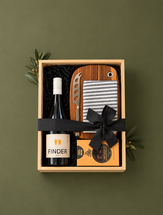A curated gift basket set in a light wood tray with black shredded filler, featuring a bottle of Finder wine, a wooden cutting board set with a striped cloth and cheese knife, and a packaged food item tied with a black ribbon.