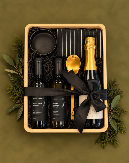 A curated gift box set in a light wood tray, featuring a bottle of Prosecco, two dark bottles, a gold spoon, a black napkin, and a black ribbon, arranged with pine branches and gold ornaments on a white background.
