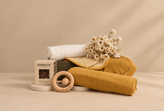 A collection of baby essentials including swaddles, bandana bibs, and a linen rattle, presented in a white magnetic box with a floral decoration.