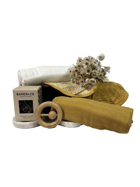A collection of baby essentials including swaddles, bandana bibs, and a linen rattle, presented in a white magnetic box with a floral decoration.