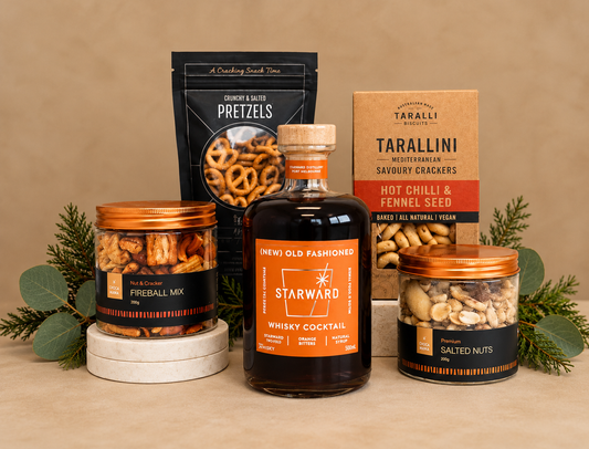 A curated gift set featuring a bottle of Starward Whiskey Cocktail, jars of Fireball Mix and Salted Nuts, a bag of Tarallini Savoury Crackers, and a bag of Chips & Lattes Pretzels, styled with a black gift bag, pine branches, and a pinecone on a white background.
