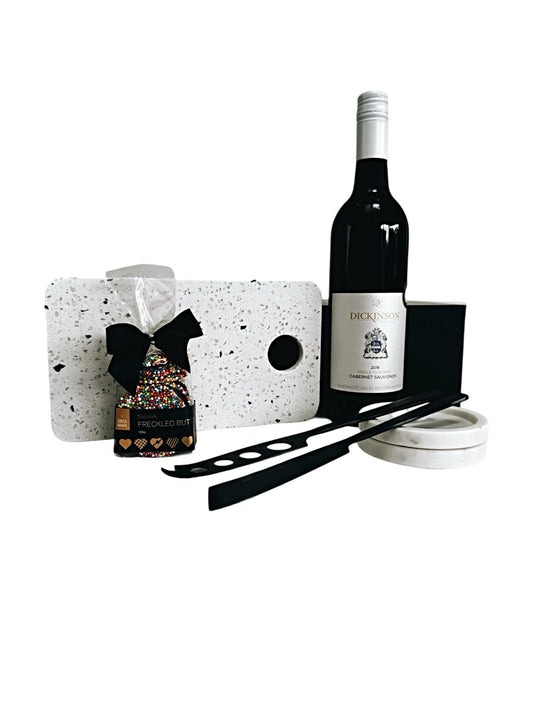 An assortment of wine and cheese accessories including a white terrazzo board, black cheese knives, a bottle of Cabernet Sauvignon, and gourmet chocolate Freckles, all presented in a magnetic gift box.