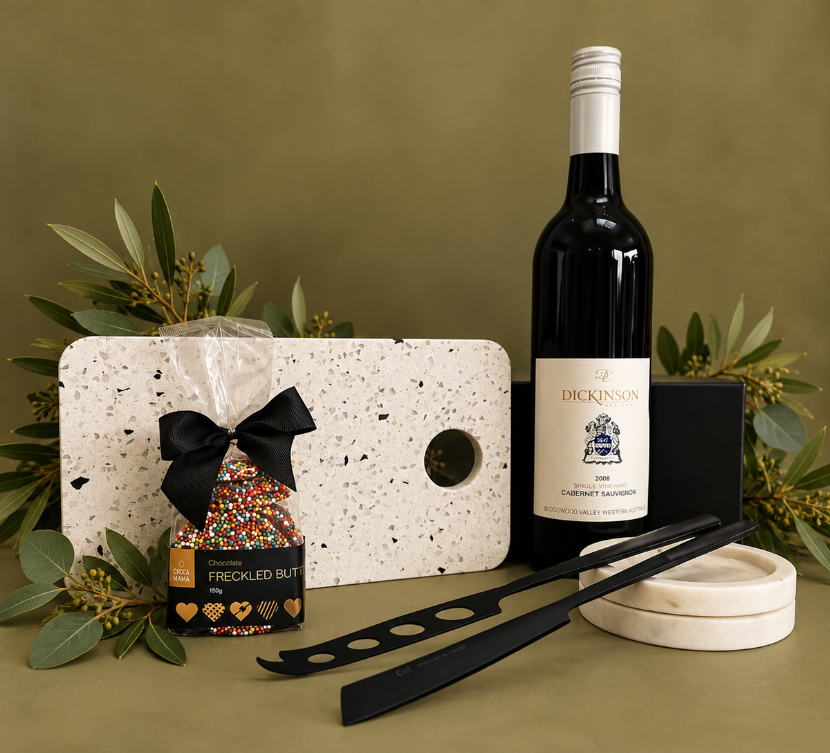 An assortment of wine and cheese accessories including a white terrazzo board, black cheese knives, a bottle of Cabernet Sauvignon, and gourmet chocolate Freckles, all presented in a magnetic gift box.