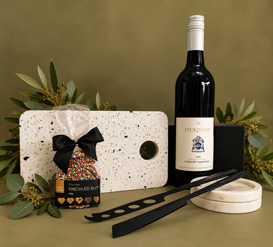 An assortment of wine and cheese accessories including a white terrazzo board, black cheese knives, a bottle of Cabernet Sauvignon, and gourmet chocolate Freckles, all presented in a magnetic gift box.